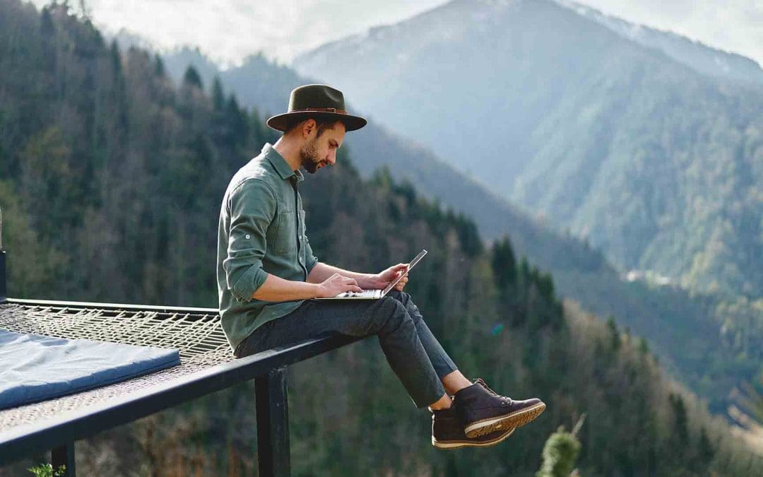 young man freelancer traveler wearing hat anywhere working online using laptop and enjoying mountains view